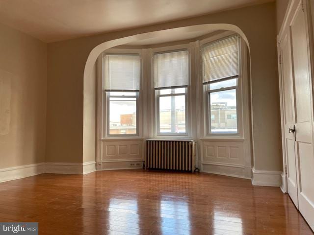 unfurnished room featuring radiator, wood finished floors, baseboards, and arched walkways