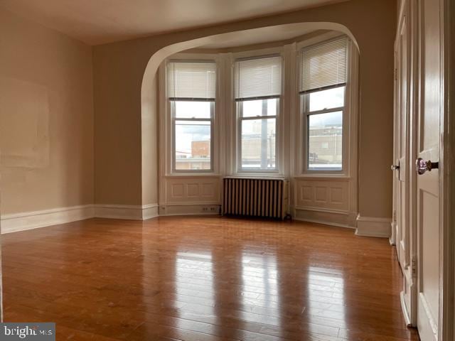 empty room featuring wood finished floors, baseboards, arched walkways, and radiator heating unit