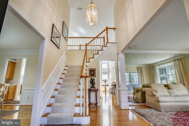staircase with a towering ceiling, wood finished floors, a chandelier, ornamental molding, and wainscoting