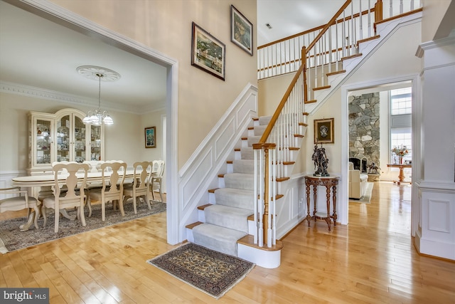staircase featuring wood-type flooring, a chandelier, ornamental molding, wainscoting, and a high ceiling