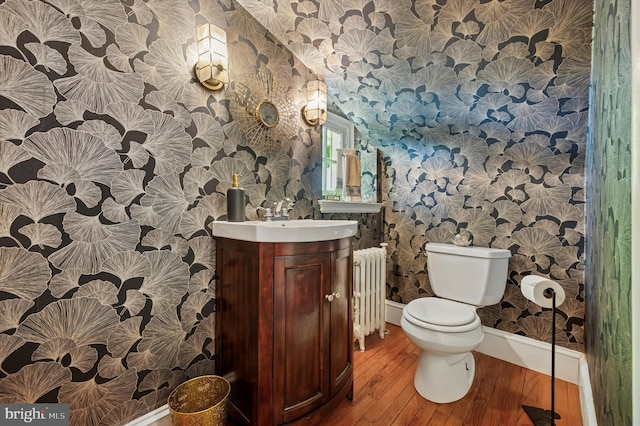 half bathroom with wallpapered walls, radiator, baseboards, wood finished floors, and toilet