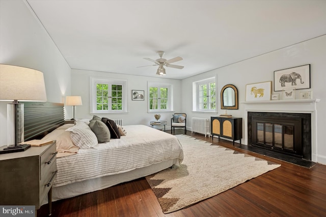 bedroom with radiator, wood finished floors, a fireplace with flush hearth, and a ceiling fan
