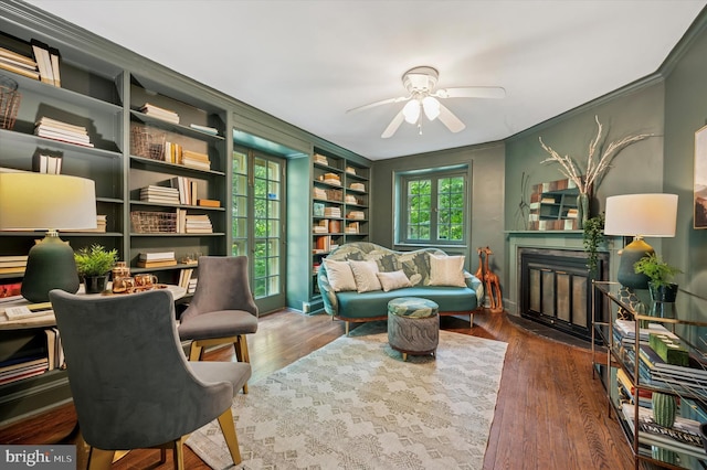 living area with ceiling fan, crown molding, wood finished floors, built in shelves, and a fireplace with flush hearth