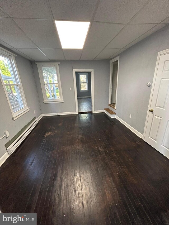 empty room featuring baseboard heating, hardwood / wood-style flooring, baseboards, and a drop ceiling