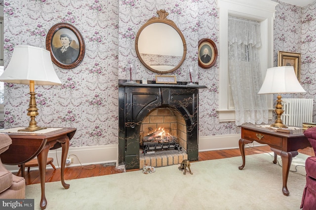 sitting room with a warm lit fireplace, radiator heating unit, wood finished floors, baseboards, and wallpapered walls