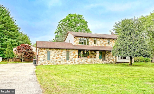 view of front facade with stone siding, a front yard, driveway, a shingled roof, and a porch