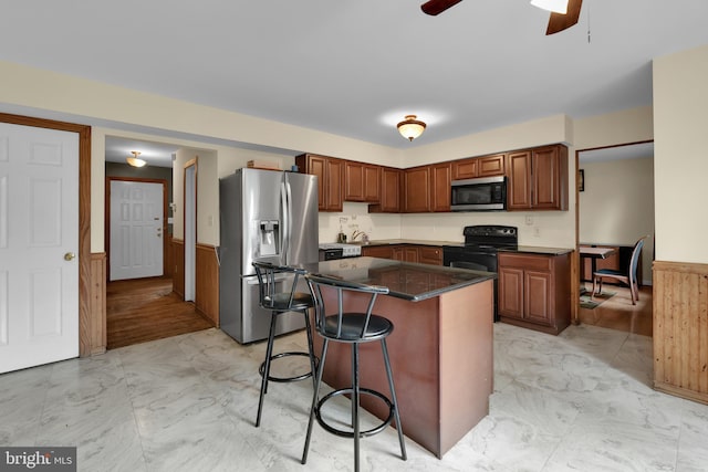 kitchen with stainless steel appliances, light marble finish flooring, wainscoting, a breakfast bar, and a ceiling fan
