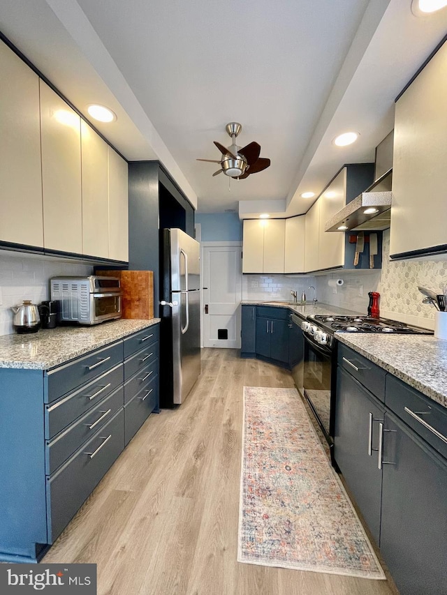 kitchen with freestanding refrigerator, black range with gas cooktop, wall chimney exhaust hood, a ceiling fan, and light wood-style floors