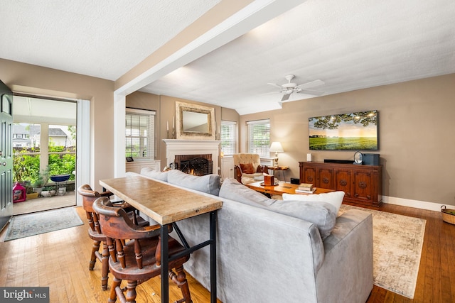 living area featuring a lit fireplace, wood finished floors, ceiling fan, baseboards, and a textured ceiling