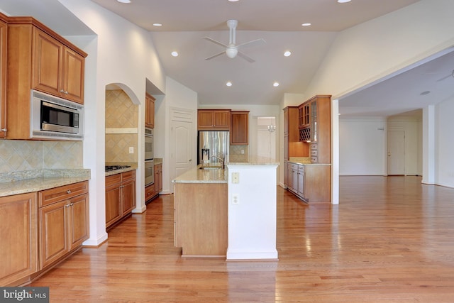 kitchen featuring appliances with stainless steel finishes, ceiling fan, light wood finished floors, a center island with sink, and light stone counters