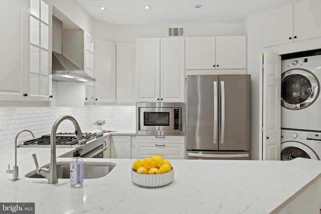 kitchen featuring appliances with stainless steel finishes, stacked washer / dryer, wall chimney range hood, white cabinets, and recessed lighting