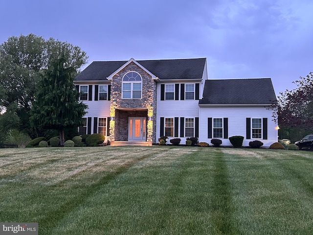 colonial inspired home with stone siding, roof with shingles, and a front lawn