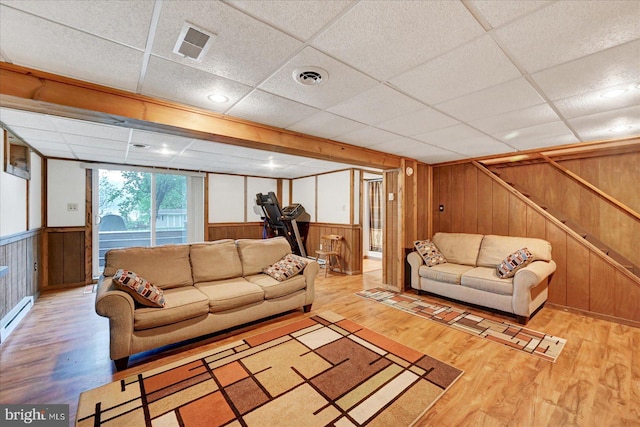 living room featuring wooden walls, wood finished floors, a paneled ceiling, baseboard heating, and stairs
