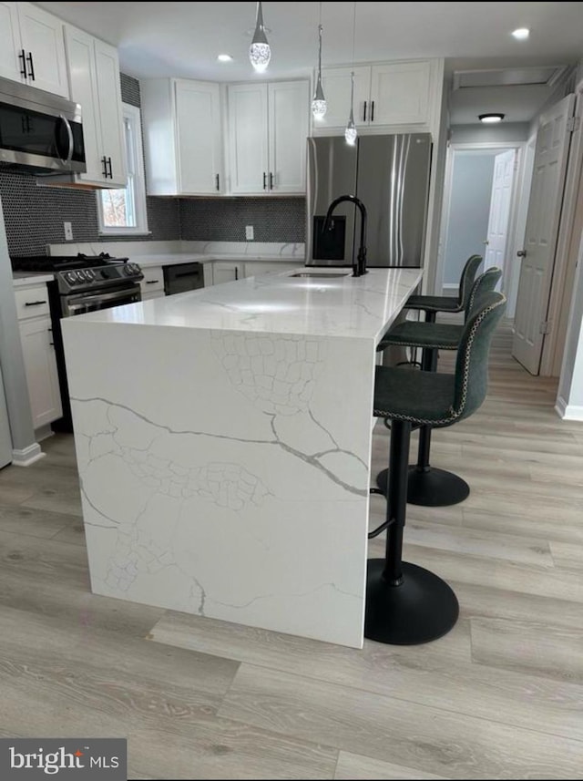 kitchen featuring light wood-type flooring, an island with sink, backsplash, white cabinets, and appliances with stainless steel finishes