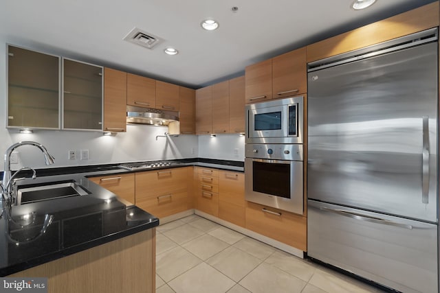 kitchen featuring built in appliances, a sink, under cabinet range hood, light tile patterned floors, and glass insert cabinets