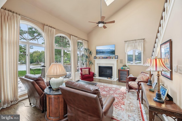 living room featuring wood finished floors, a glass covered fireplace, a ceiling fan, and high vaulted ceiling