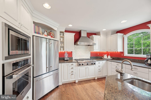 kitchen with stainless steel appliances, wall chimney range hood, a sink, white cabinets, and dark stone countertops