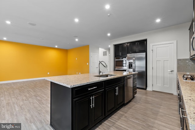kitchen with a sink, appliances with stainless steel finishes, dark cabinets, a center island with sink, and light wood-type flooring