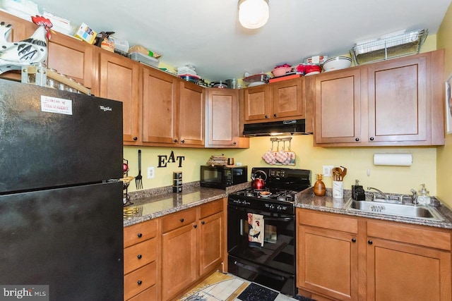 kitchen featuring black appliances, a sink, under cabinet range hood, brown cabinetry, and dark stone countertops