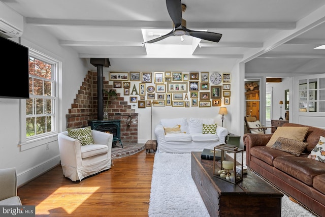 living room featuring wood finished floors, a wood stove, ceiling fan, beamed ceiling, and a wall unit AC