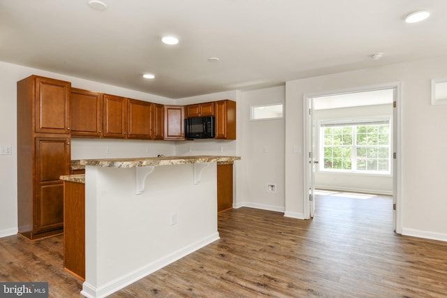kitchen with black microwave, a kitchen bar, brown cabinetry, dark wood-type flooring, and recessed lighting