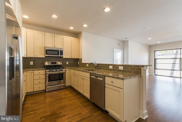 kitchen with stone countertops, dark wood-style flooring, a peninsula, decorative backsplash, and appliances with stainless steel finishes