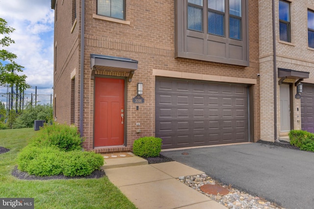view of exterior entry with a garage, brick siding, driveway, and central air condition unit
