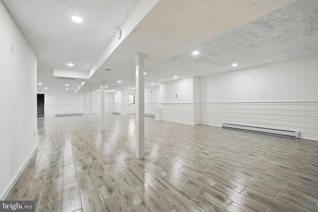 below grade area featuring a textured ceiling, a wainscoted wall, baseboard heating, and wood finished floors
