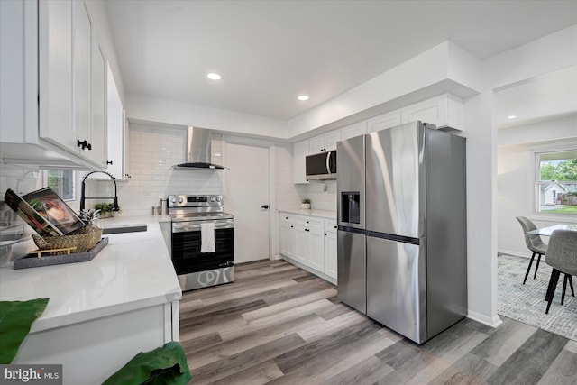 kitchen featuring appliances with stainless steel finishes, wall chimney exhaust hood, light wood-style flooring, white cabinets, and tasteful backsplash
