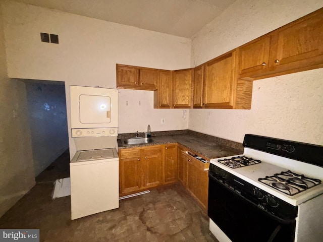 kitchen featuring gas range oven, a sink, brown cabinets, dark countertops, and stacked washer / drying machine