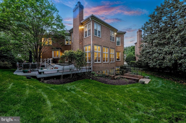 back of house at dusk featuring a chimney, a lawn, a deck, and brick siding