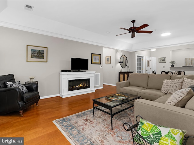 living room featuring ceiling fan, light wood-type flooring, baseboards, a glass covered fireplace, and crown molding