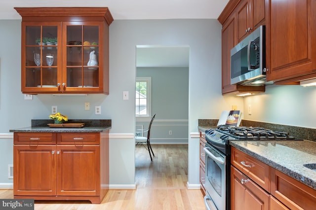 kitchen featuring stainless steel appliances, dark stone counters, light wood-style flooring, glass insert cabinets, and baseboards