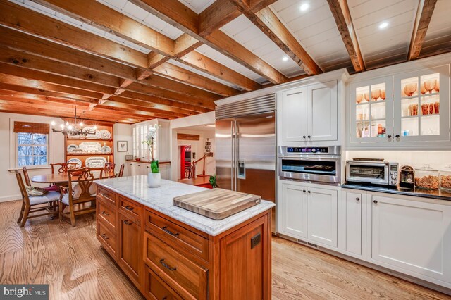 kitchen featuring stainless steel appliances, light wood finished floors, light stone counters, white cabinetry, and beam ceiling