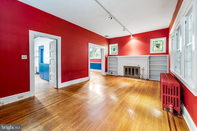 unfurnished living room featuring radiator heating unit, wood-type flooring, track lighting, baseboards, and a brick fireplace