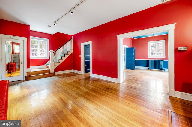 unfurnished living room featuring radiator, track lighting, wood-type flooring, stairway, and baseboards