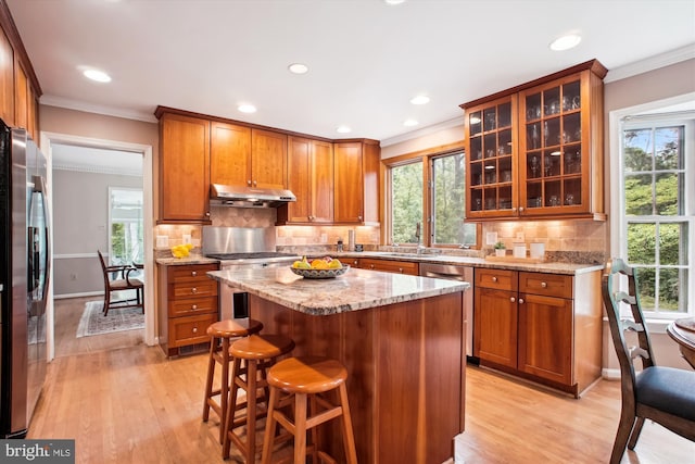 kitchen featuring appliances with stainless steel finishes, under cabinet range hood, crown molding, brown cabinetry, and light wood-type flooring
