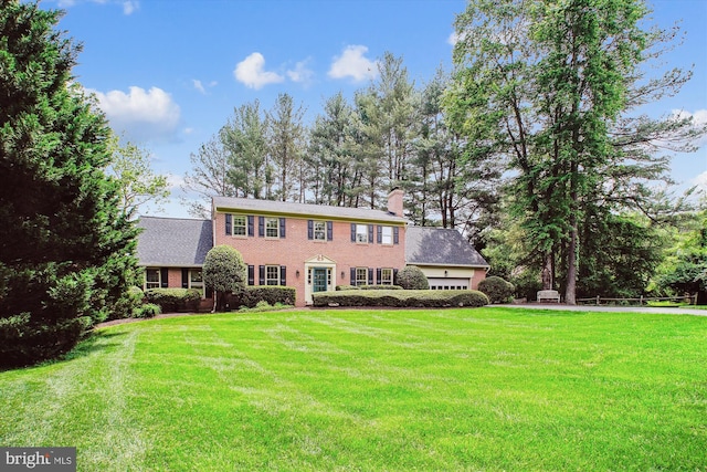 colonial house with an attached garage, a front yard, a chimney, and brick siding