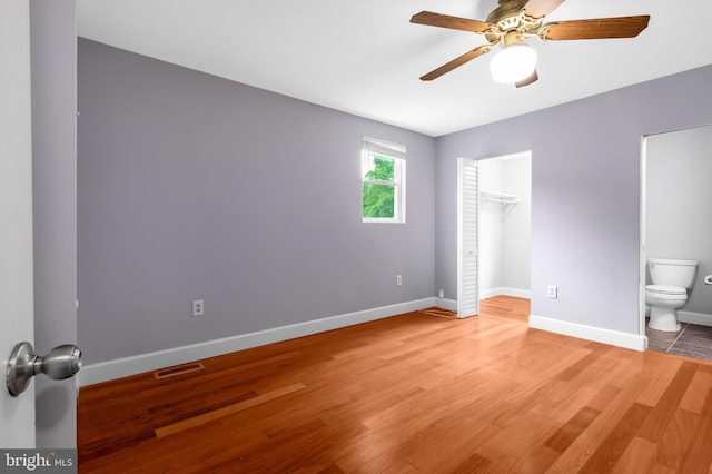 unfurnished bedroom featuring baseboards, light wood finished floors, ensuite bath, a spacious closet, and a ceiling fan