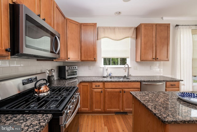 kitchen featuring stainless steel appliances, a sink, light wood-type flooring, dark stone countertops, and tasteful backsplash