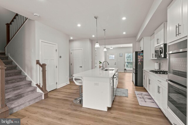 kitchen with a sink, a center island with sink, stainless steel appliances, white cabinets, and light wood-type flooring