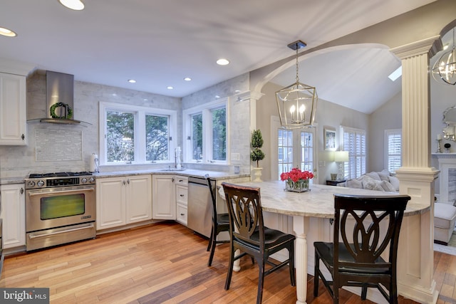 kitchen featuring a peninsula, stainless steel appliances, wall chimney range hood, ornate columns, and light wood-type flooring