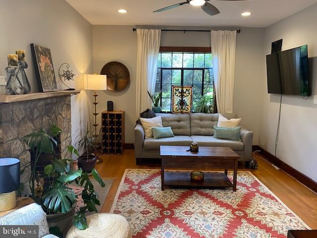 living area with wood finished floors, ceiling fan, baseboards, a stone fireplace, and recessed lighting