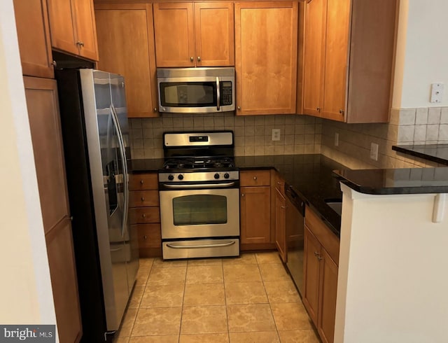 kitchen featuring brown cabinetry, tasteful backsplash, light tile patterned floors, dark countertops, and appliances with stainless steel finishes