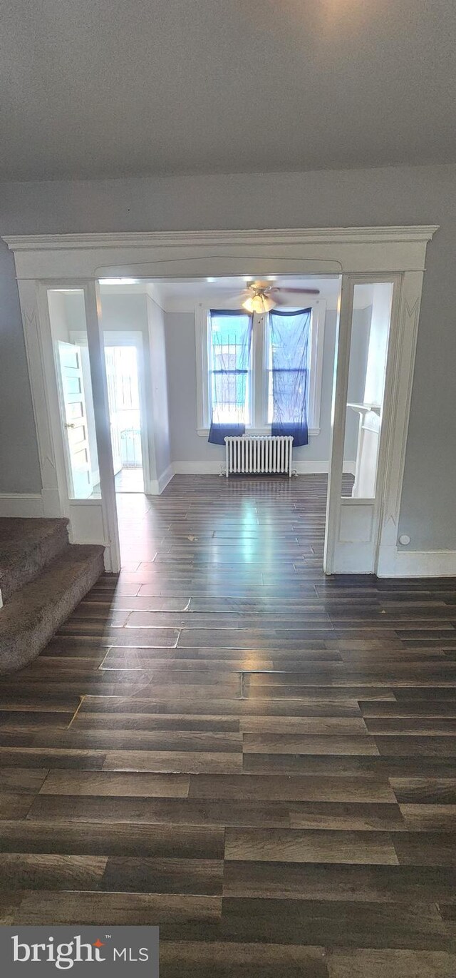 empty room featuring radiator, dark wood-style floors, baseboards, stairs, and a ceiling fan