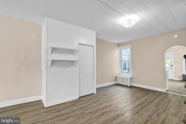 unfurnished living room featuring arched walkways, radiator heating unit, wood finished floors, and baseboards