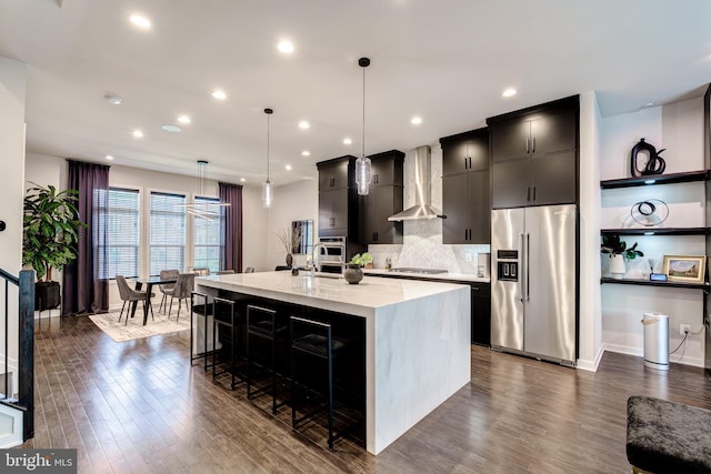 kitchen featuring appliances with stainless steel finishes, wall chimney range hood, a kitchen island with sink, dark wood finished floors, and recessed lighting