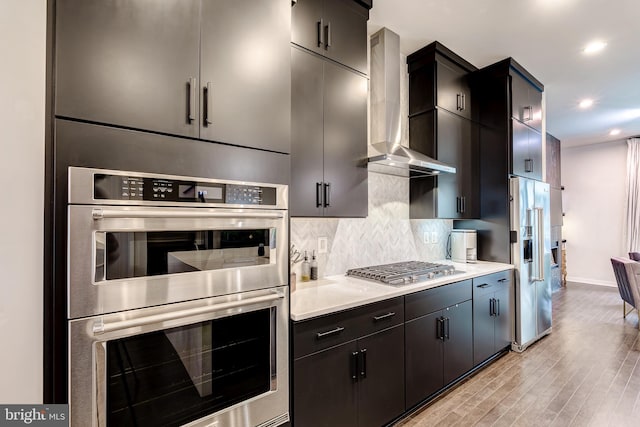kitchen featuring appliances with stainless steel finishes, wall chimney exhaust hood, baseboards, decorative backsplash, and light wood-type flooring