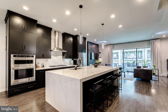 kitchen featuring wall chimney exhaust hood, dark cabinets, a large island, stainless steel appliances, and decorative backsplash