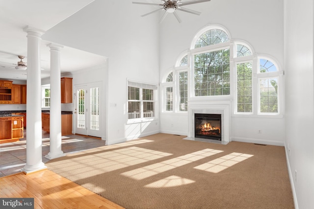 unfurnished living room featuring ceiling fan, a high ceiling, ornate columns, a fireplace with flush hearth, and healthy amount of natural light